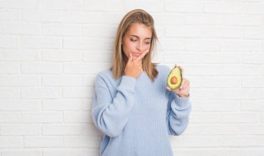 Güzel genç kadın hakkında soru, çok karışık bir fikir taze avokado ciddi yüz düşünme yemek beyaz tuğla duvarın üzerinden