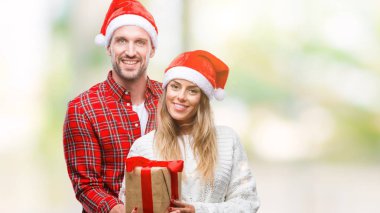 Noel şapka ve izole arka plan üzerinde duran ve diş gösterilen kendine güvenen bir gülümsemeyle gülümseyerek mutlu bir yüz ile mevcut tutarak aşık genç çift