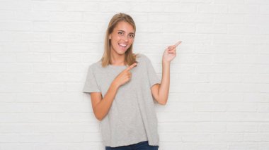 Güzel genç kadın gülümseyerek ve iki elleri ve parmakları ile tarafına işaret kamera bakıyor beyaz tuğla duvarın üzerinden.