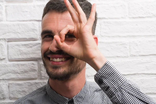 Genç yetişkin adam mutlu yüz el parmaklarının arasından seyir göz üzerinde ok işaretiyle yapıyor gülümseyen ile beyaz tuğla duvarı ayakta