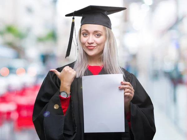 指していると幸せそうな顔を笑顔で側まで親指で示す分離の背景上の学位を保持する大学院の制服を着て若いブロンドの女性 — ストック写真