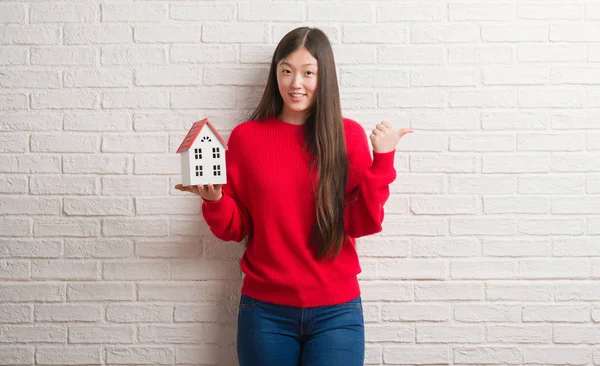 Genç Çinli gerçek durum Aracısı kadın işaret ve başparmak kadar mutlu yüz gülümseyen tarafı ile gösterilen tuğla duvar holding ev
