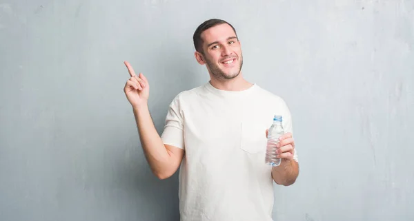 側の手と指で指す非常に幸せな水のボトルを保持している灰色のグランジの壁を越えて若い白人男 — ストック写真