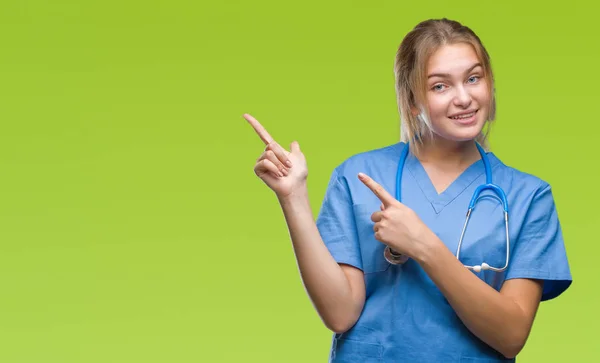 年轻的高加索医生妇女穿着外科医生制服在孤立的背景微笑着 看着相机指向镜头 用两只手和手指指向一边 — 图库照片