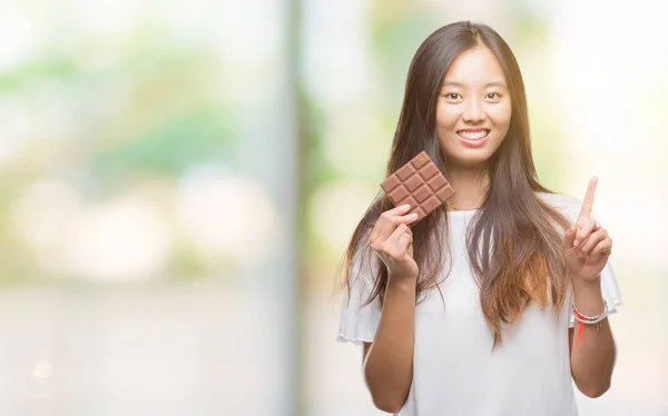 Young asian woman eating chocolate bar over isolated background surprised with an idea or question pointing finger with happy face, number one