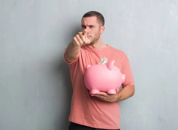 Joven Hombre Caucásico Sobre Pared Grunge Gris Sosteniendo Alcancía Señalando — Foto de Stock