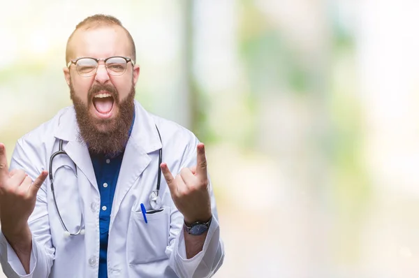 Young caucasian doctor man wearing medical white coat over isolated background shouting with crazy expression doing rock symbol with hands up. Music star. Heavy concept.