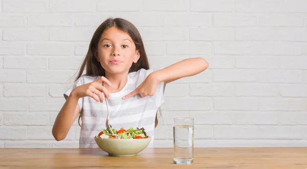 年轻的西班牙裔孩子坐在桌子上吃健康沙拉与惊讶的脸指向自己的手指 — 图库照片
