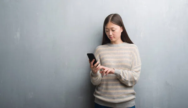 灰色墙上的年轻中国妇女看着智能手机与自信的表情 认真思考 — 图库照片