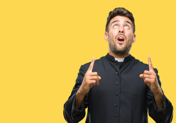 Young catholic christian priest man over isolated background amazed and surprised looking up and pointing with fingers and raised arms.