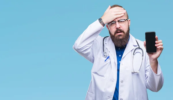 Young doctor man showing smartphone screen over isolated background stressed with hand on head, shocked with shame and surprise face, angry and frustrated. Fear and upset for mistake.