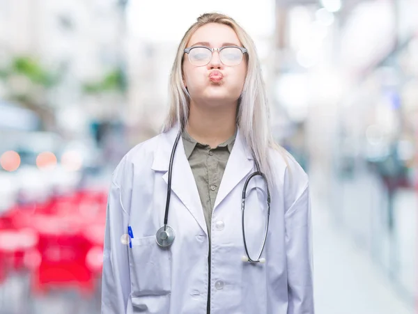 Young blonde doctor woman over isolated background puffing cheeks with funny face. Mouth inflated with air, crazy expression.