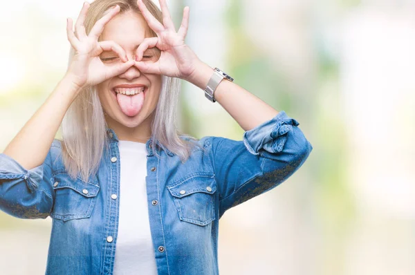 Young blonde woman over isolated background doing ok gesture like binoculars sticking tongue out, eyes looking through fingers. Crazy expression.