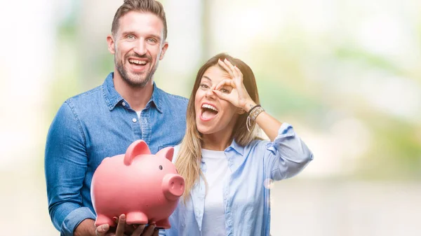 Young couple in love holding piggy bank over isolated background with happy face smiling doing ok sign with hand on eye looking through fingers