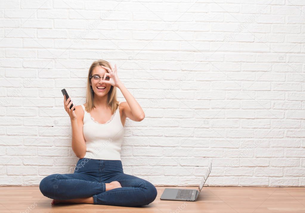 Beautiful young woman sitting on the floor texting using smartphone and laptop with happy face smiling doing ok sign with hand on eye looking through fingers