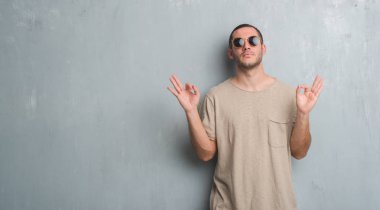 Güneş gözlüğü takıyor gri grunge duvarın üzerinden beyaz delikanlı rahatla ve gülümseyen gözlerle kapalı parmak jestle meditasyon yapıyor. Yoga kavramı.
