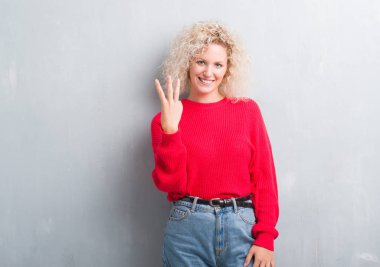 Gösterilen ve parmakları ile işaret grunge gri arka plan üzerinde Kıvırcık saçlı sarışın genç kadın kendinden emin ve mutlu gülümseyerek iken üçüncü.