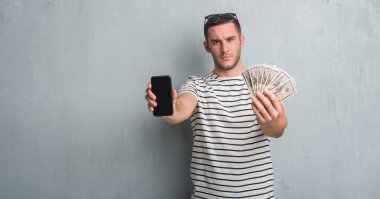 Beyaz delikanlı holding sorunu nedeniyle dolar ve smarphone şüpheci ve sinir, papatyanın üzgün gri grunge duvarın üzerinden. Negatif kişi.