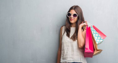 Grunge gri duvar renkli alışveriş torbaları kendine güvenen bir ifade ile acımak yüz ciddi düşünüyorum tutan genç esmer kadın