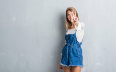 Zafer işareti yapıyor parmak gösterilen kamera seyir gülümseyen grunge gri duvar üzerinde duran güzel genç kadın. İki numaralı.