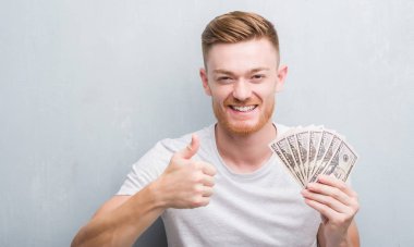 Genç Kızıl saçlı adam dolar ile mutlu büyük holding gri grunge duvarın üzerinden gülümseme ok işareti, başparmak ile parmak, mükemmel işareti yapıyor