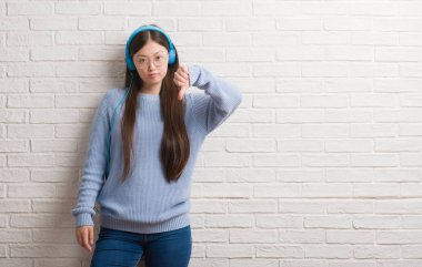 Kızgın yüz, eksi işareti aşağı antipati başparmak ile gösterilen, ret kavramı ile kulaklıklar giyen tuğla duvarın üzerinden Genç Çinli kadın