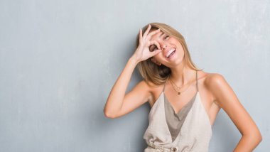 Güzel genç kadın mutlu yüz el parmaklarının arasından seyir göz üzerinde ok işaretiyle yapıyor gülümseyen grunge gri duvardan ayakta