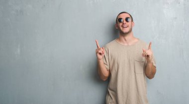 Beyaz delikanlı güneş gözlüğü takıyor gri grunge duvarın üzerinden hayran ve yukarıya bakıyor ve parmakları ile işaret ve silah kaldırdı şaşırttı.