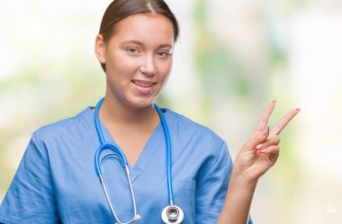 Mutlu yüz zafer işareti yaparken kameraya göz kırpıyor ile gülümseyen izole arka plan üzerinde tıbbi üniforma giyen genç beyaz doktor kadın. İki numaralı.