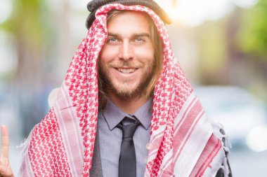 Genç yakışıklı Arap adam uzun saçlı ve parmak gülümseyen süre bir numara ile kendinden emin ve mutlu yukarıyı gösteren izole arka plan üzerinde keffiyeh giyiyor.