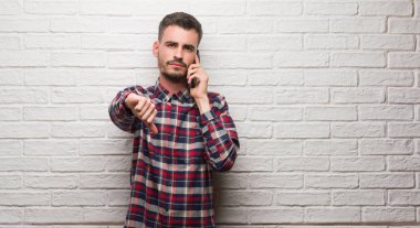 Kızgın yüz, eksi işareti aşağı antipati başparmak ile gösterilen, ret kavramı ile beyaz tuğla duvarın üzerinden ayakta telefonda konuşan genç yetişkin adam