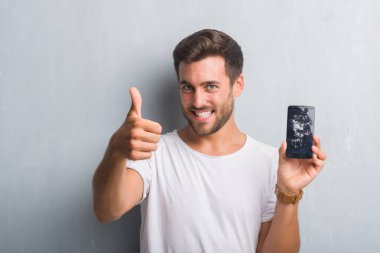 Yakışıklı genç adam akıllı telefon ekran ile mutlu büyük kırık gösterilen gri grunge duvarın üzerinden gülümseme ok işareti, başparmak ile parmak, mükemmel işareti yapıyor