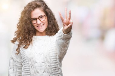 Güzel esmer kıvırcık saçlı Genç kız giyiyor kış kazak gösterilen ve parmakları ile işaret izole arka plan üzerinde üçüncü ise kendinden emin ve mutlu gülümseyerek.