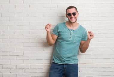 Duygu tezahürat Genç beyaz adam ayakta beyaz tuğla duvarı gurur çığlık ve zafer ve başarı çok heyecanlı, kutluyor güneş gözlüğü takıyor