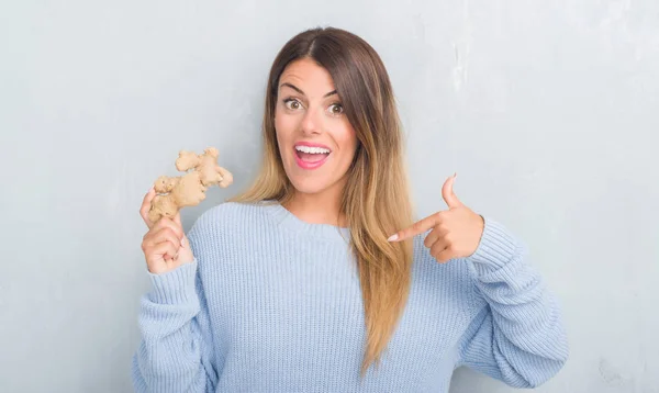 Young adult woman over grey grunge wall holding fresh ginger vegatable with surprise face pointing finger to himself