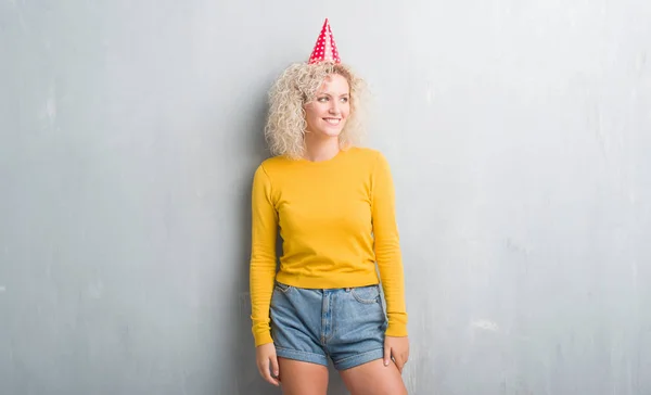若いブロンドの女性グランジに灰色の壁着て誕生日帽子よそ見側に笑顔で顔に自然な表現 自信を持って笑ってください — ストック写真
