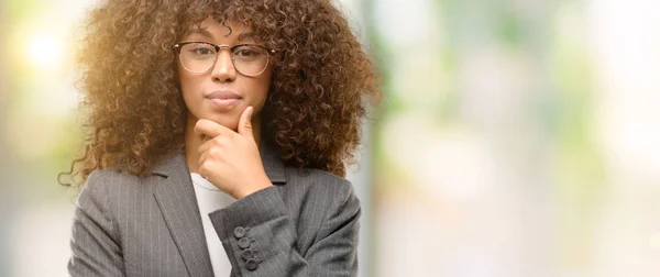 African american business woman wearing glasses looking confident at the camera with smile with crossed arms and hand raised on chin. Thinking positive.