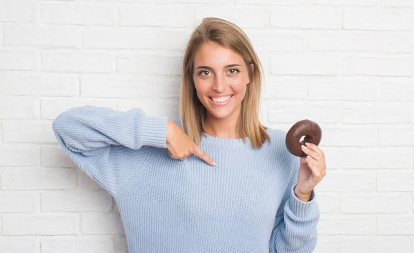 自分自身に驚き顔ポインティング指でチョコレート ドーナツを食べる白いレンガの壁に美しい若い女性 — ストック写真