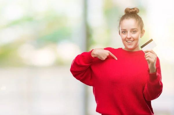 Young blonde woman holding credit card with surprise face pointing finger to himself