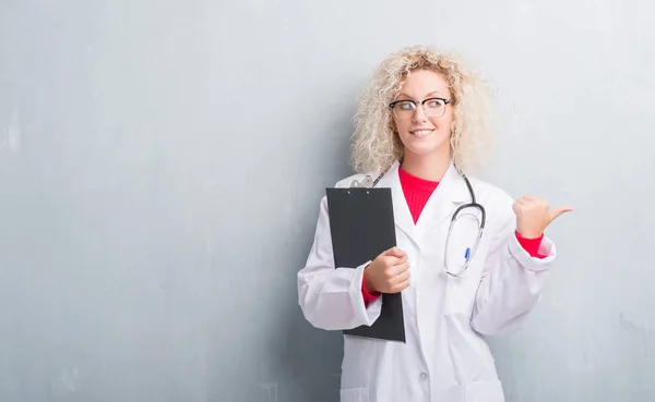 Young blonde doctor woman over grunge grey wall holding clipboard pointing and showing with thumb up to the side with happy face smiling