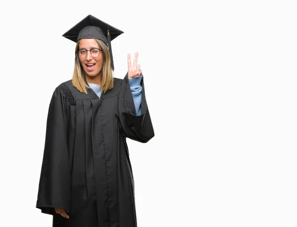 着た若い美しい女性は 勝利のサインを行うカメラでウィンクしている幸せそうな顔を浮かべて隔離された背景に制服を卒業しました — ストック写真