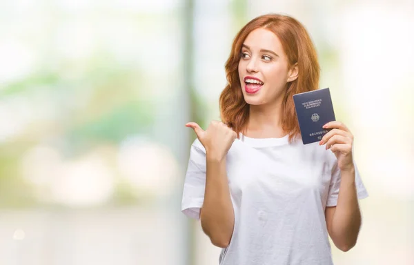 拿着德国护照的年轻美丽的妇女在隔绝的背景指向和显示与拇指由边与愉快的面孔微笑 — 图库照片