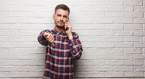 Kızgın yüz, eksi işareti aşağı antipati başparmak ile gösterilen, ret kavramı ile beyaz tuğla duvarın üzerinden ayakta telefonda konuşan genç yetişkin adam