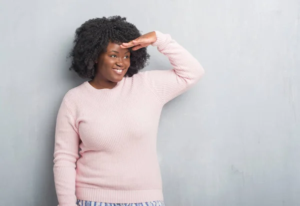 Young african american plus size woman over grey grunge wall wearing winter sweater very happy and smiling looking far away with hand over head. Searching concept.