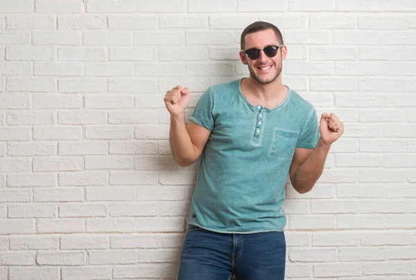 Duygu tezahürat Genç beyaz adam ayakta beyaz tuğla duvarı gurur çığlık ve zafer ve başarı çok heyecanlı, kutluyor güneş gözlüğü takıyor