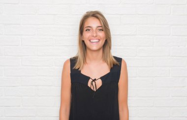 Güzel genç kadın ayakta ve diş gösterilen kendine güvenen bir gülümsemeyle gülümseyerek mutlu bir yüz ile beyaz tuğla duvarın üzerinden