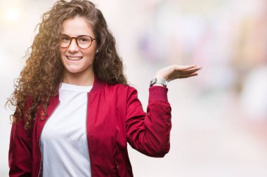 Ceket ve gözlük neşeli sunulması ve kameraya bakarak avucunun ile işaret gülümseyen izole arka plan üzerinde giyen güzel esmer kıvırcık saçlı Genç kız.