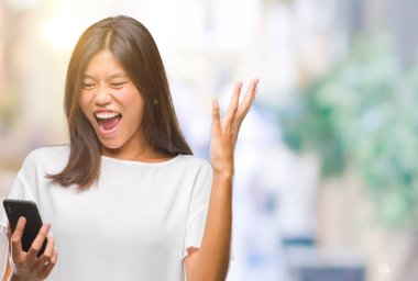 Genç Asyalı kadın manifatura üzerinden Smartphone kullanarak büyük gülümseme ile çığlık zaferini kutluyor arka plan çok mutlu ve heyecanlı, kazanan ifade izole ve ellerini kaldırdı