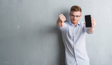 Genç Kızıl saçlı adam gri grunge duvar boş ekran smartphone kızgın yüzü, eksi işareti aşağı antipati başparmak ile gösterilen, ret kavramı ile gösterilen