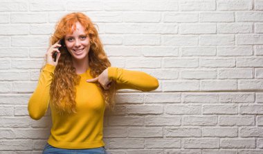 Kendisi için sürpriz yüz işaret parmağı ile telefonda konuşurken tuğla duvarın üzerinden duran genç Kızıl saçlı kadın